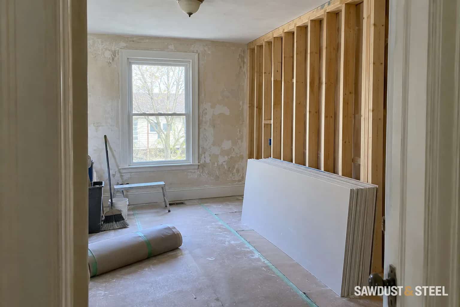 A room with timber partition framing, stacked plasterboard, and protected flooring