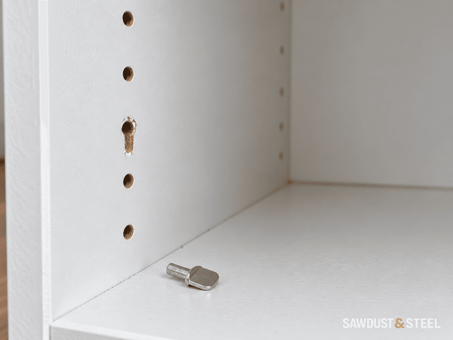A chipped shelf-support hole in a white cabinet side, with the loose metal pin on the empty cabinet bottom