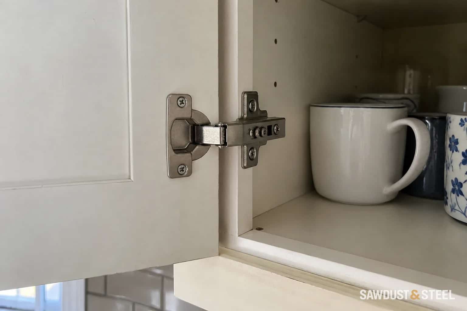 Concealed hinge joining an open white cabinet door to a cabinet containing mugs