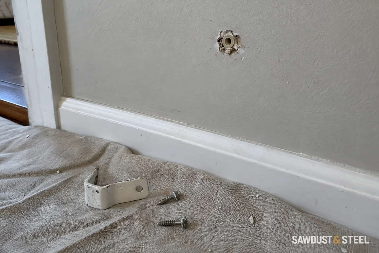 Damaged wall-anchor hole above a loose metal bracket and screws on a drop cloth