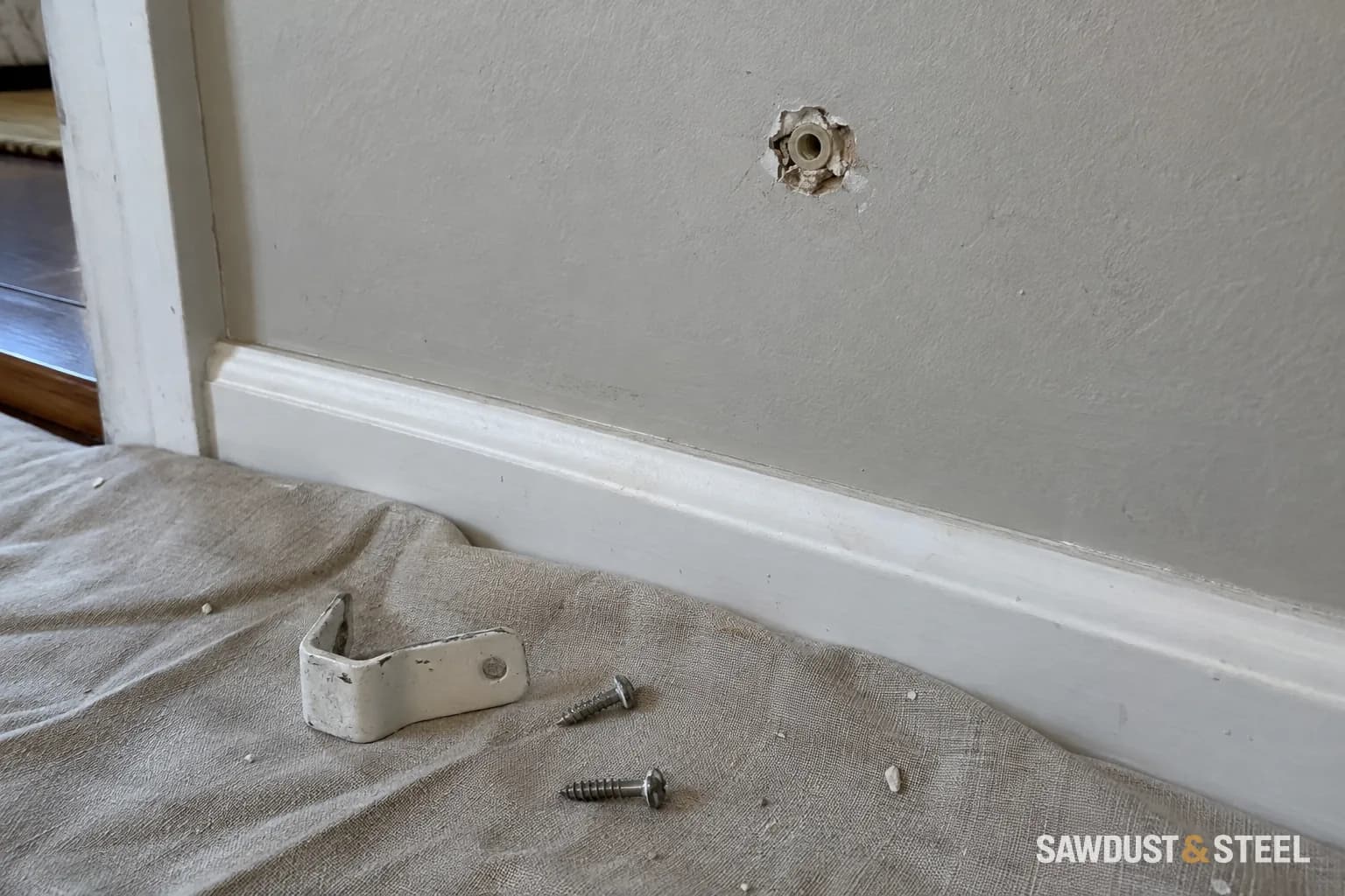 Damaged wall-anchor hole above a loose metal bracket and screws on a drop cloth