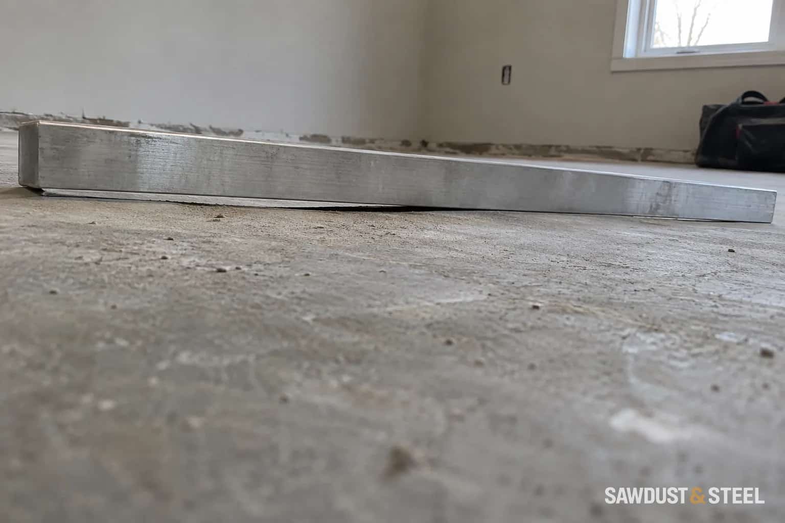Long metal straightedge resting on a bare concrete floor with a small gap visible beneath the middle