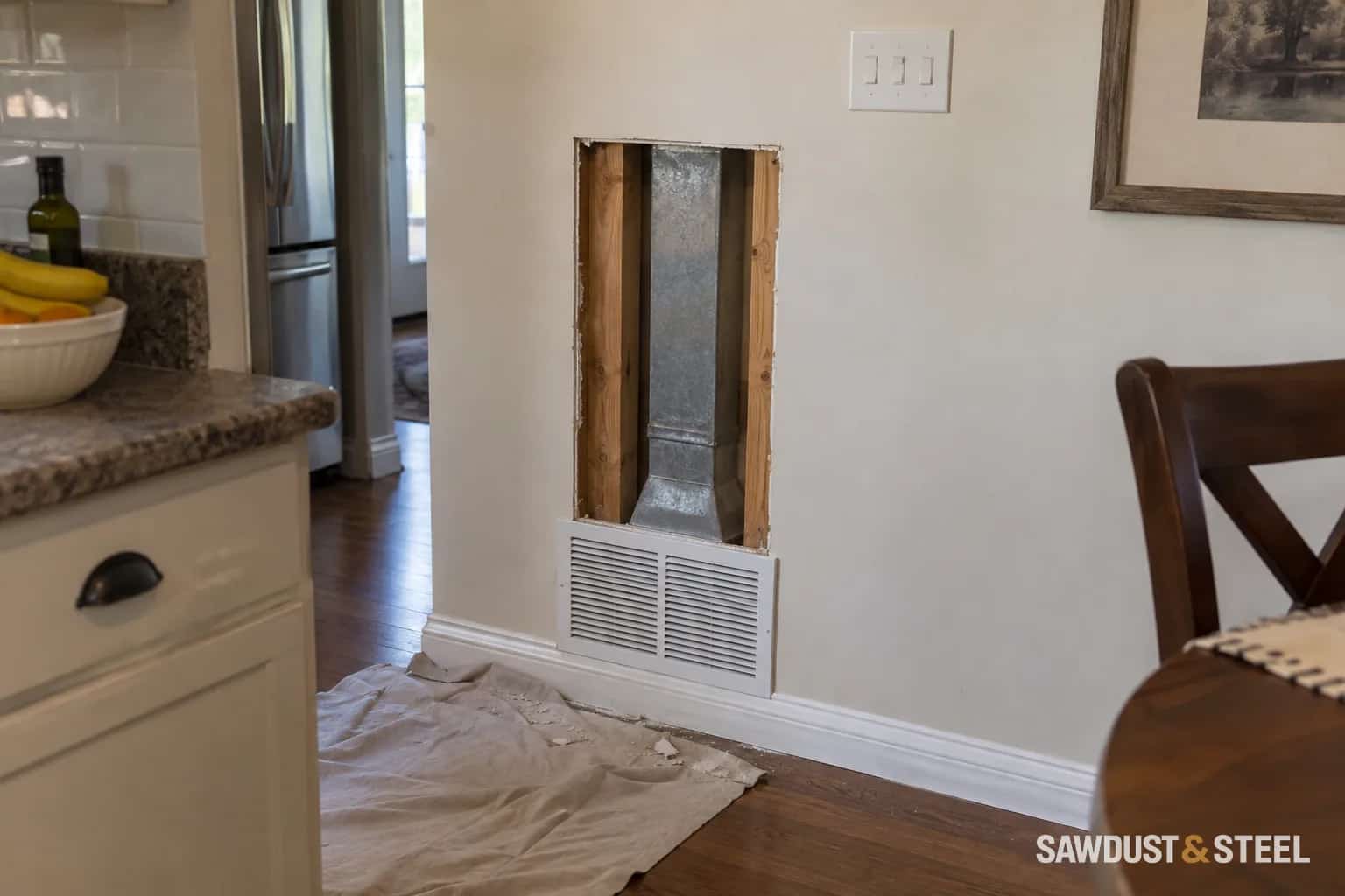 Opened drywall showing a rectangular metal duct between intact studs above a low wall grille.