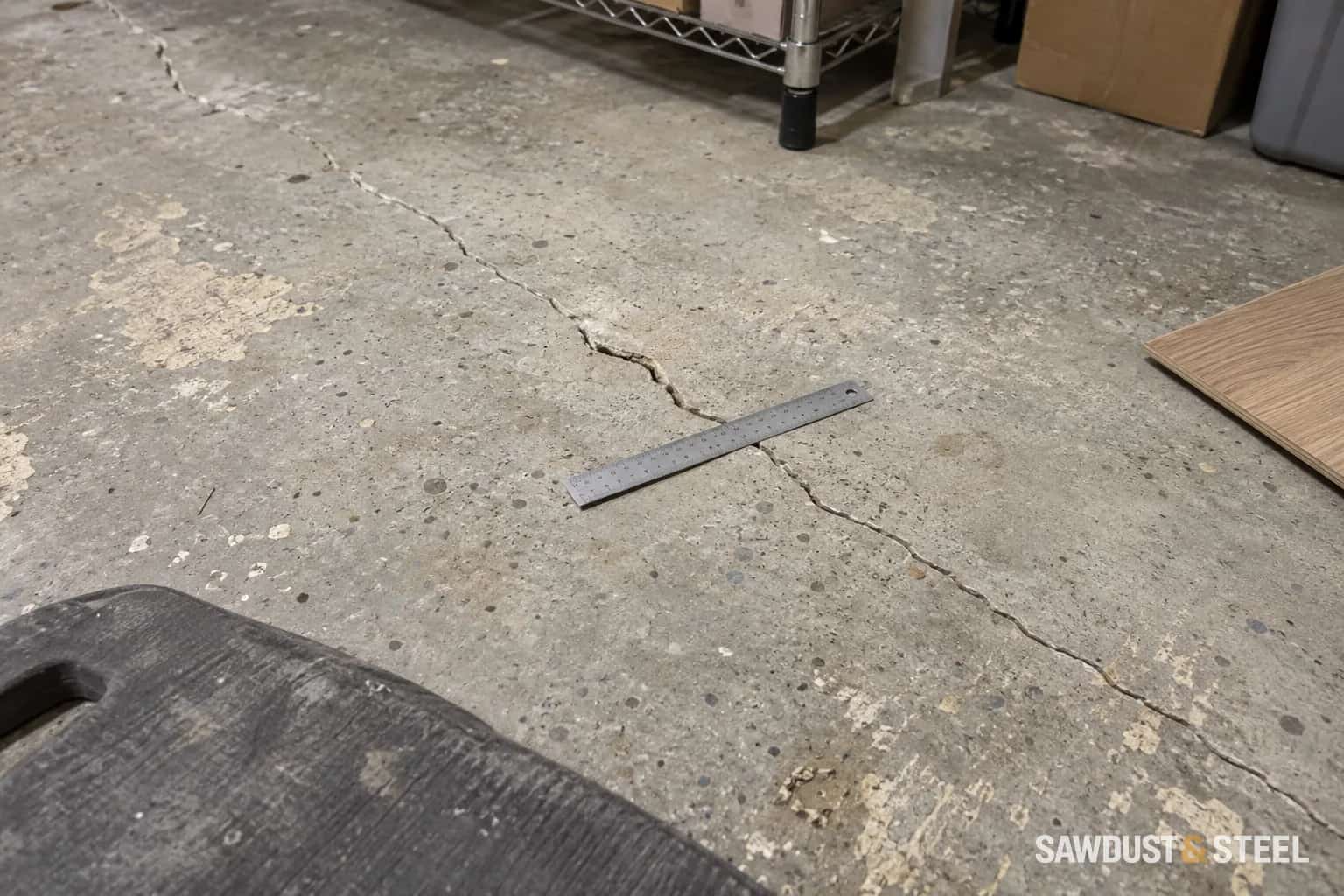 Ruler laid across an exposed basement slab crack beside a flooring sample