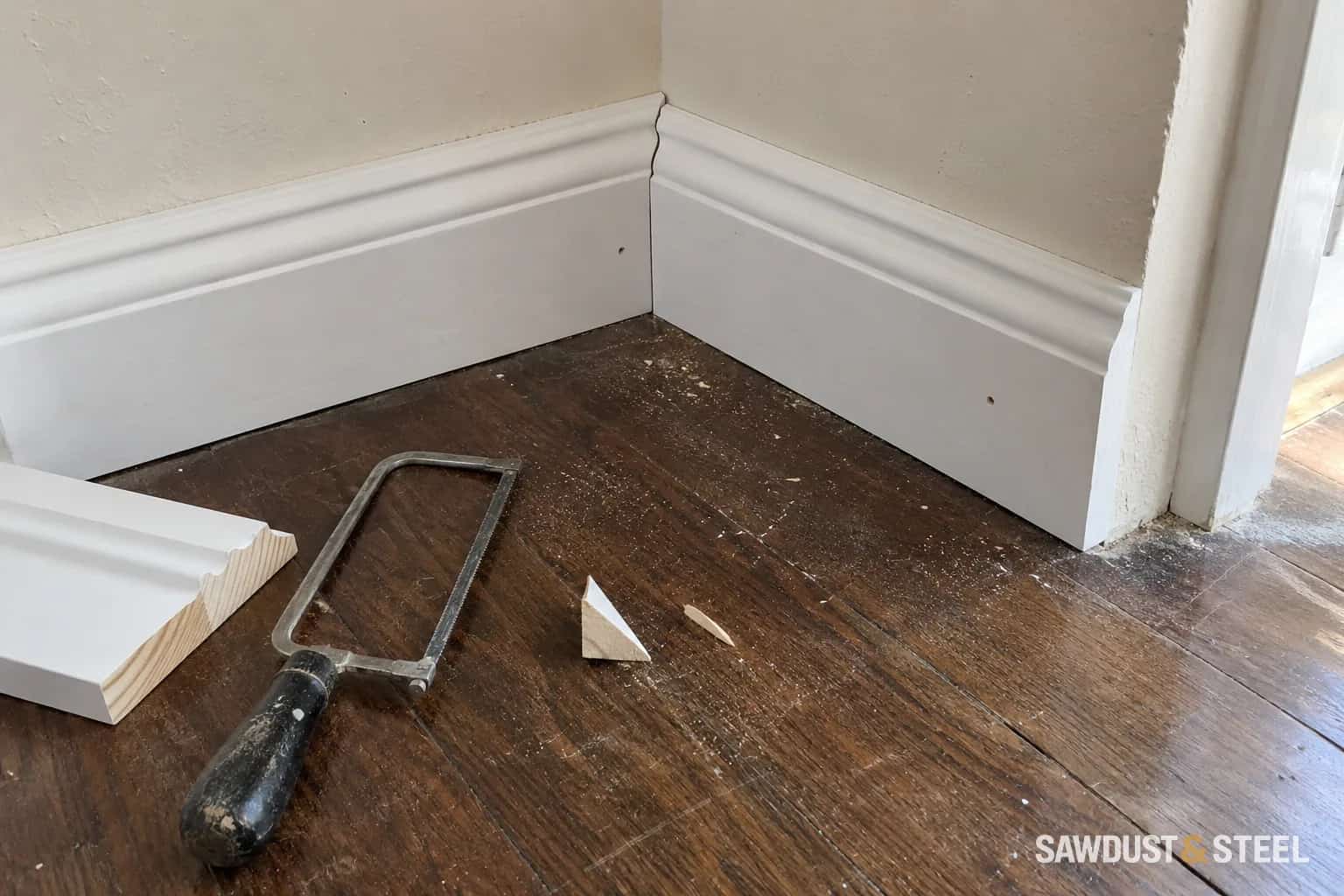 Shaped primed baseboard meeting at an inside corner with a matching offcut and coping saw on the floor