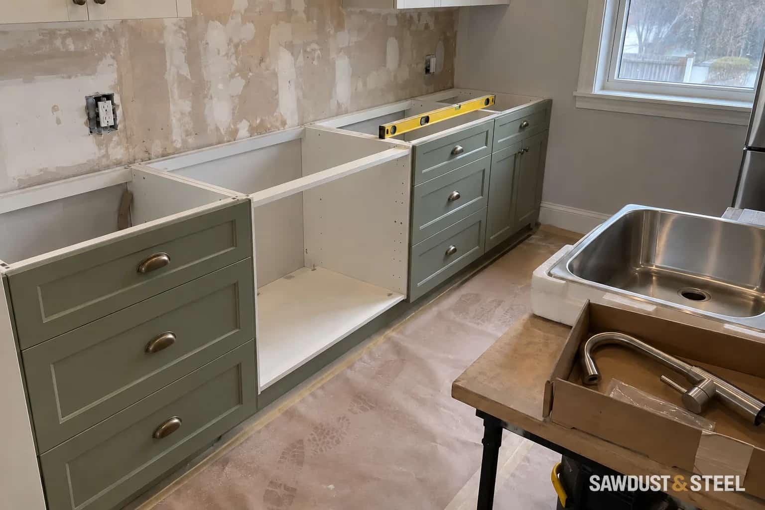 Sage base cabinets awaiting countertops, with a sink and boxed tap on the nearby work table
