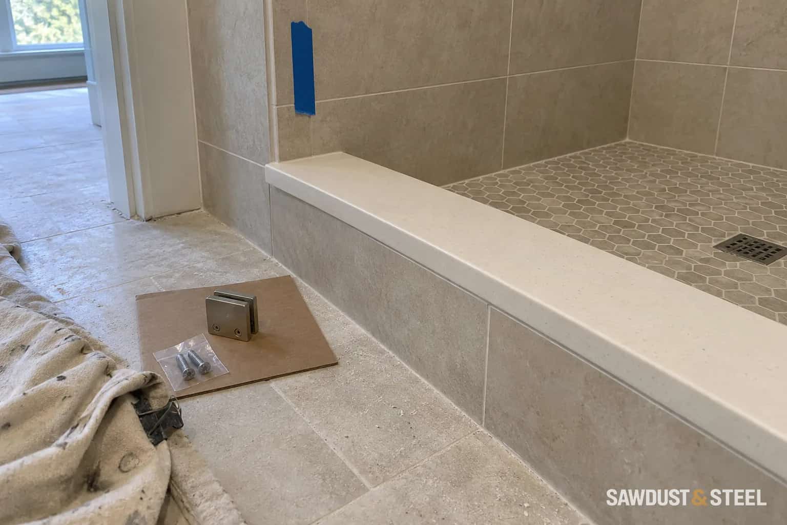 Tiled shower curb with an intact stone cap, a loose glass bracket, and bagged fasteners on cardboard