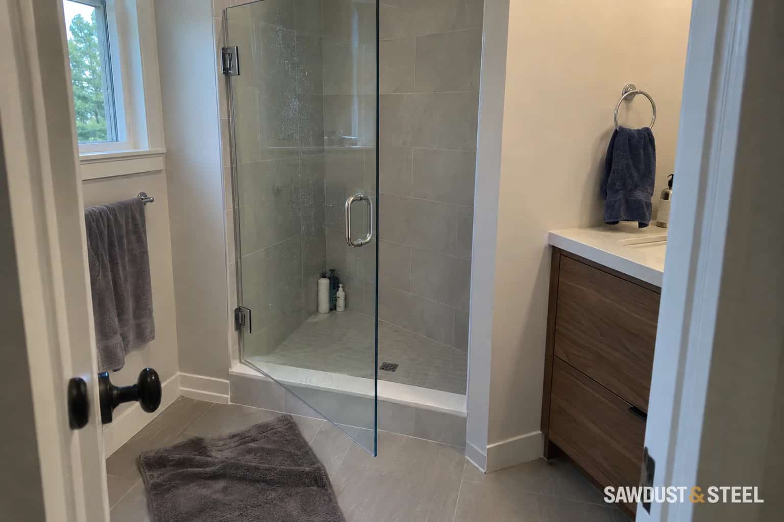 Glass shower enclosure beside a wood vanity in a tiled bathroom