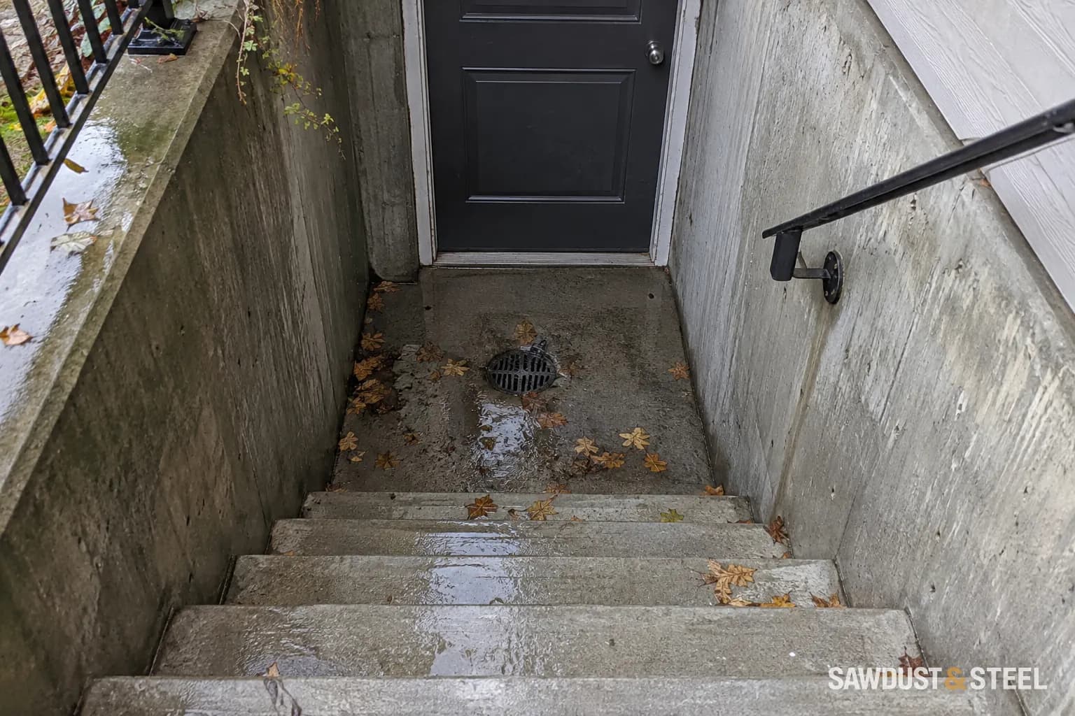 Wet concrete basement stairwell with a grated drain and fallen leaves on the landing.