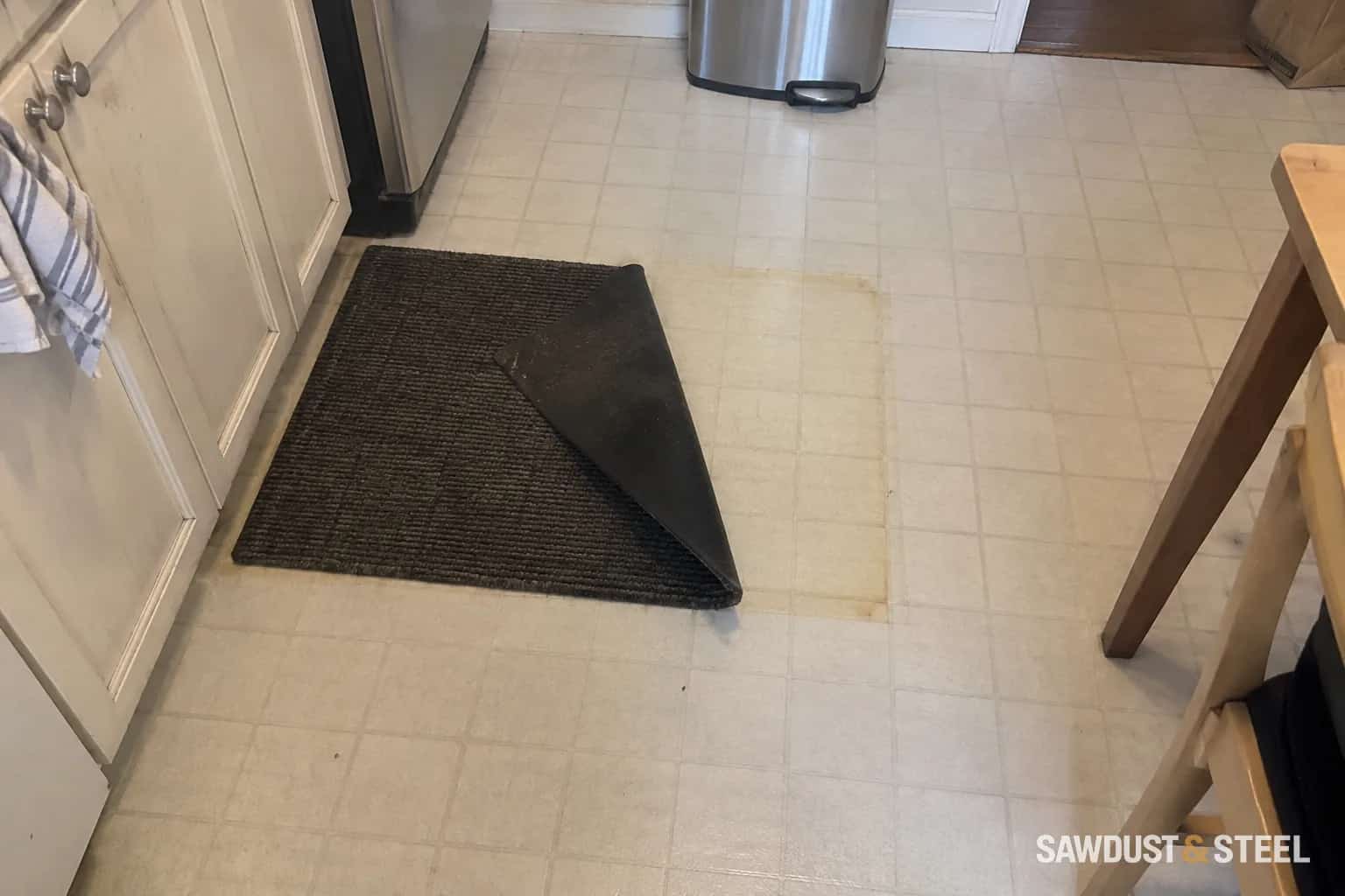 A kitchen mat folded back over a yellowed patch of pale vinyl flooring.