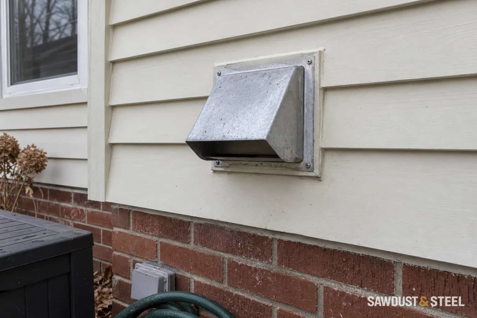 Metal exhaust wall cap on cream siding above a brick foundation