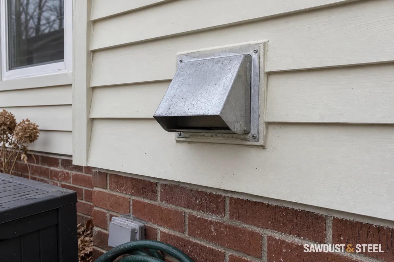 Metal exhaust wall cap on cream siding above a brick foundation