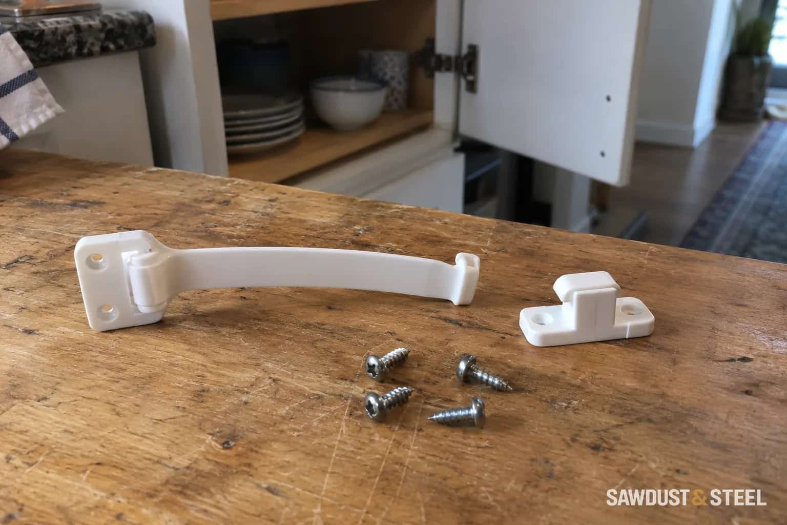 Uninstalled white cabinet safety latch, separate catch and four screws on a wooden workbench