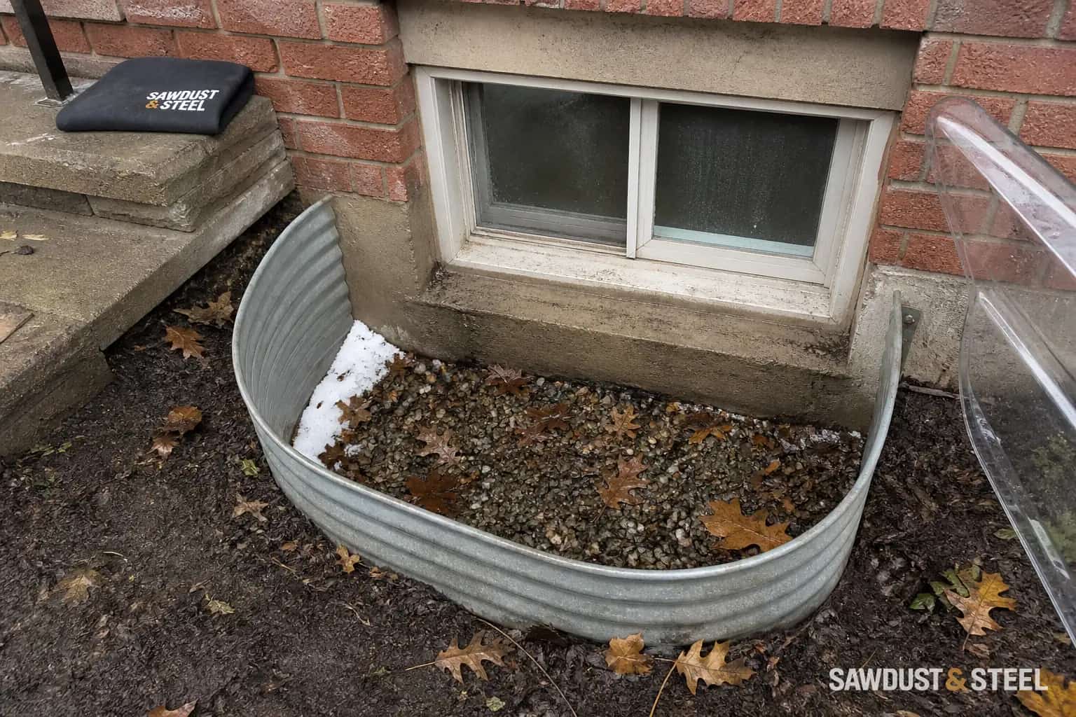 Editorial view of a basement window well with damp gravel and autumn leaves beside a brick house